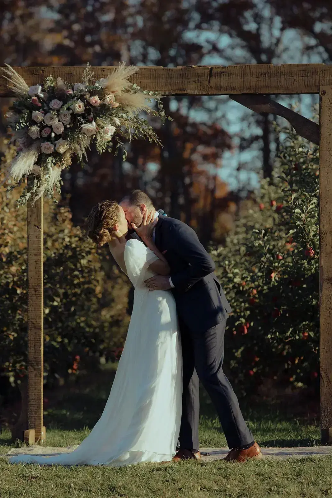 First kiss under the wooden arch with the orchard behind them